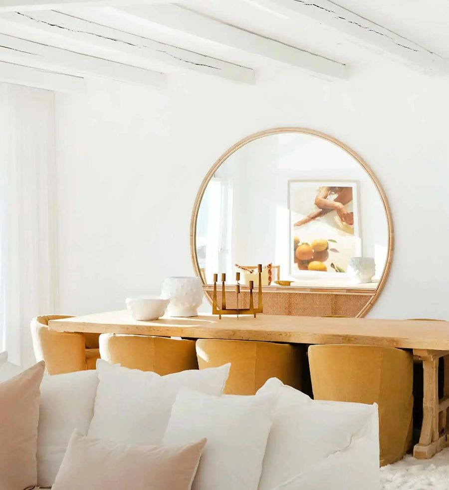 Lifestyle shot of a minimalist dining room featuring a large round mirror reflecting an art print. The art print, titled "Pool Time II", depicts a close-up of a person's legs and torso partially submerged in water, with warm light reflecting on the skin. Below the person, a still life of oranges and lemons is visible. The room has white walls and exposed white ceiling beams, a light wood dining table, and mustard yellow upholstered chairs. A white sofa with white and blush pink pillows is in the foreground.
