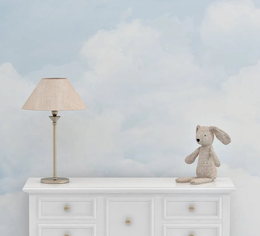 Nursery:
Peel and stick wallpaper featuring a repeating pattern of soft, fluffy white clouds against a pale blue sky, creating a serene and dreamy atmosphere. The wallpaper is displayed in a nursery setting above a white dresser with gold knobs, with a beige table lamp and a small, light brown stuffed bunny sitting on the dresser.