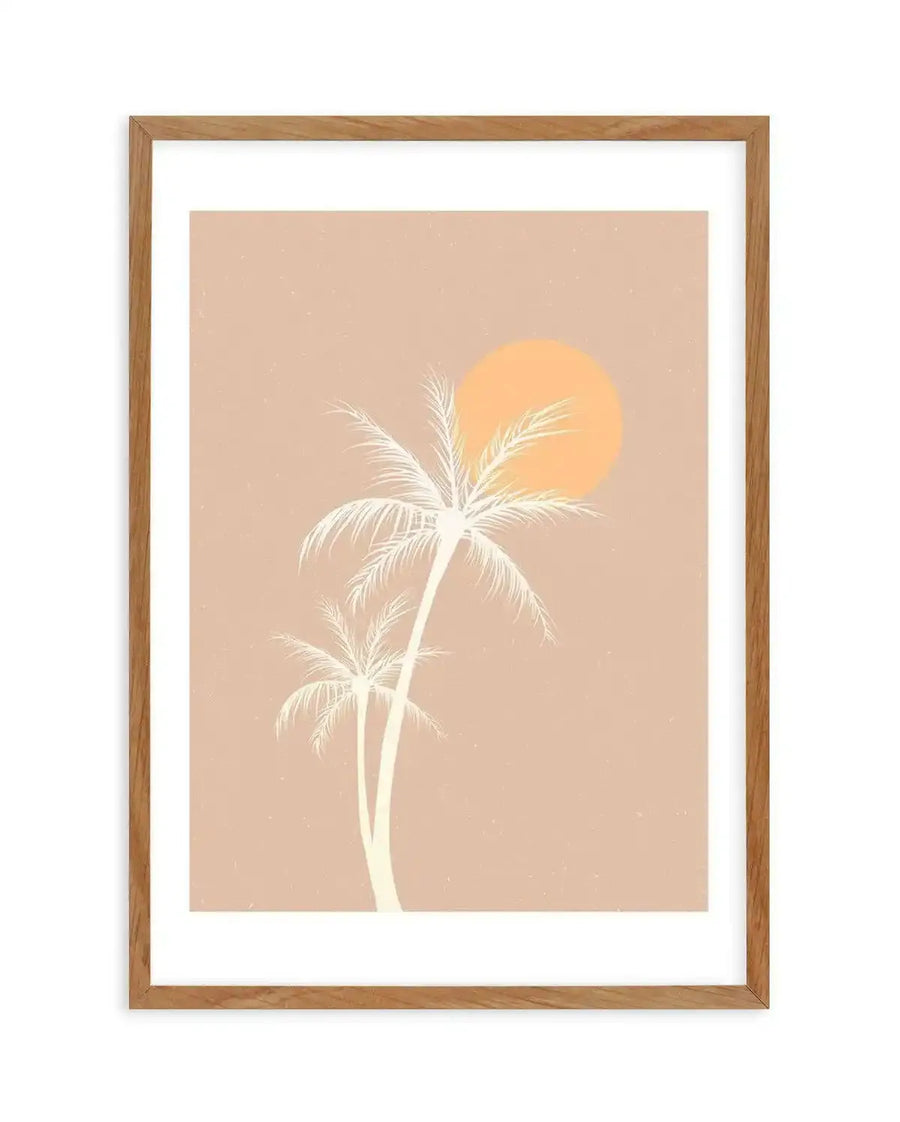 Art print featuring two white palm trees silhouetted against a warm peach-pink background with a large, soft orange sun in the upper right, printed on fine art paper and displayed in a natural oak frame with white border.