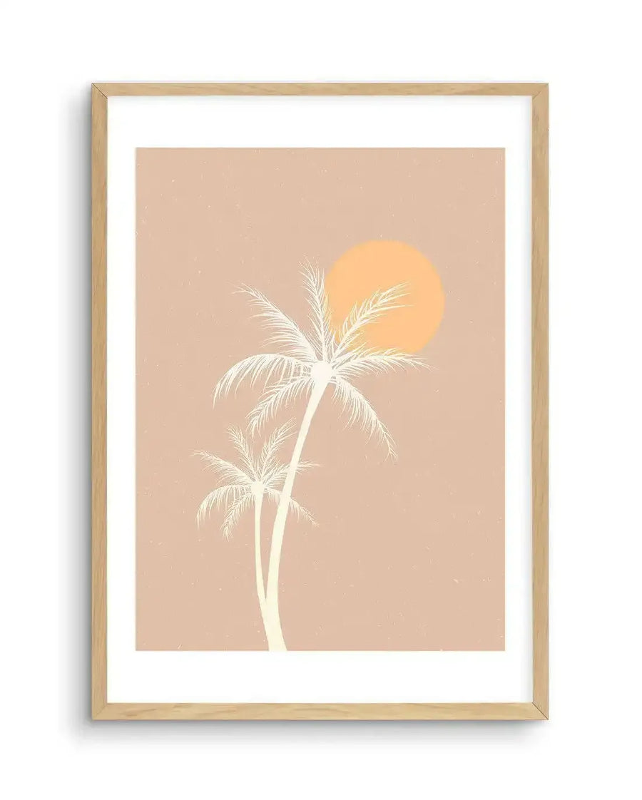 Art print featuring an illustration of two palm trees in a minimalist style against a warm, dusty pink background with a large, bright orange sun. The palm trees are depicted in a light cream colour with fine details on the fronds, and the overall composition evokes a retro, sunset beach scene. The print is displayed in a natural oak frame with a white border.