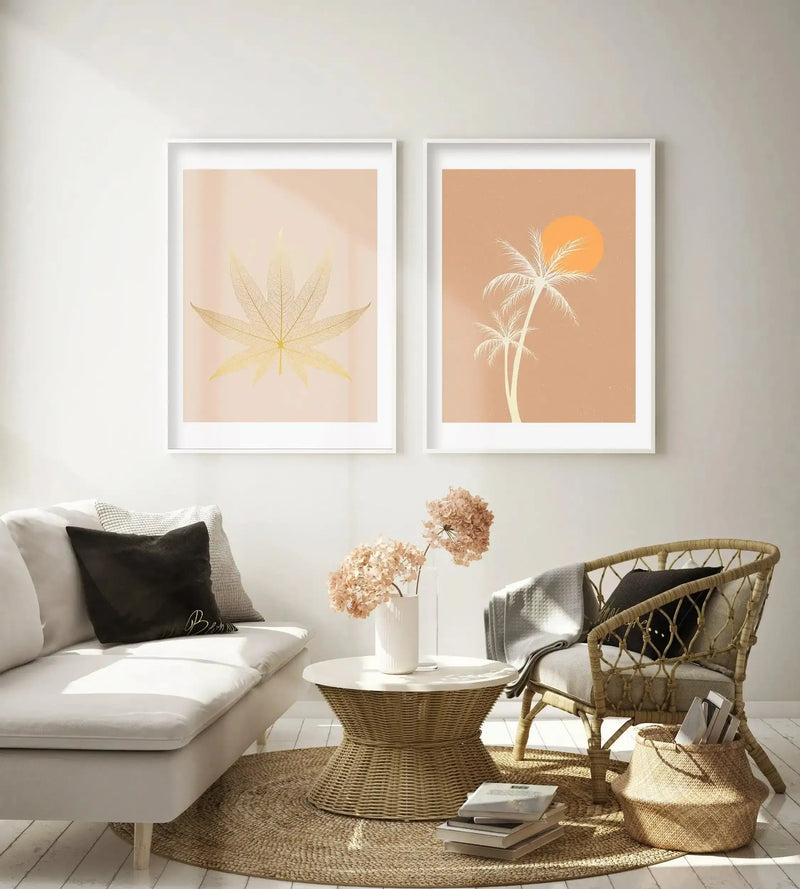 Lifestyle shot of two framed art prints displayed above a curved white sofa in a minimalist living room. The left print features a golden skeletal leaf on a pale peach background, and the right print features white palm trees and an orange sun on a warm terracotta background. The room includes a woven coffee table, a wicker armchair, and dried flowers in a white vase.