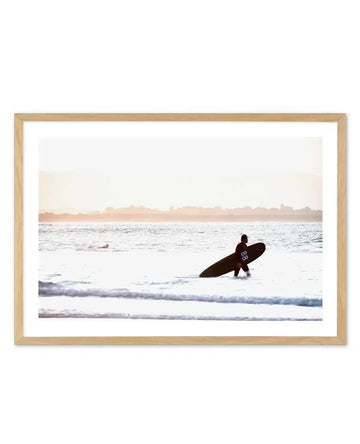 Framed photography art print of a coastal scene at sunset with a natural wood frame and white border. A surfer wearing a black wetsuit with the number "88" on the back holds a dark surfboard and wades into the ocean, with gentle waves breaking around them. Another surfer is visible further out in the water. The sky is a soft, hazy orange and pink, with a faint silhouette of land on the horizon.