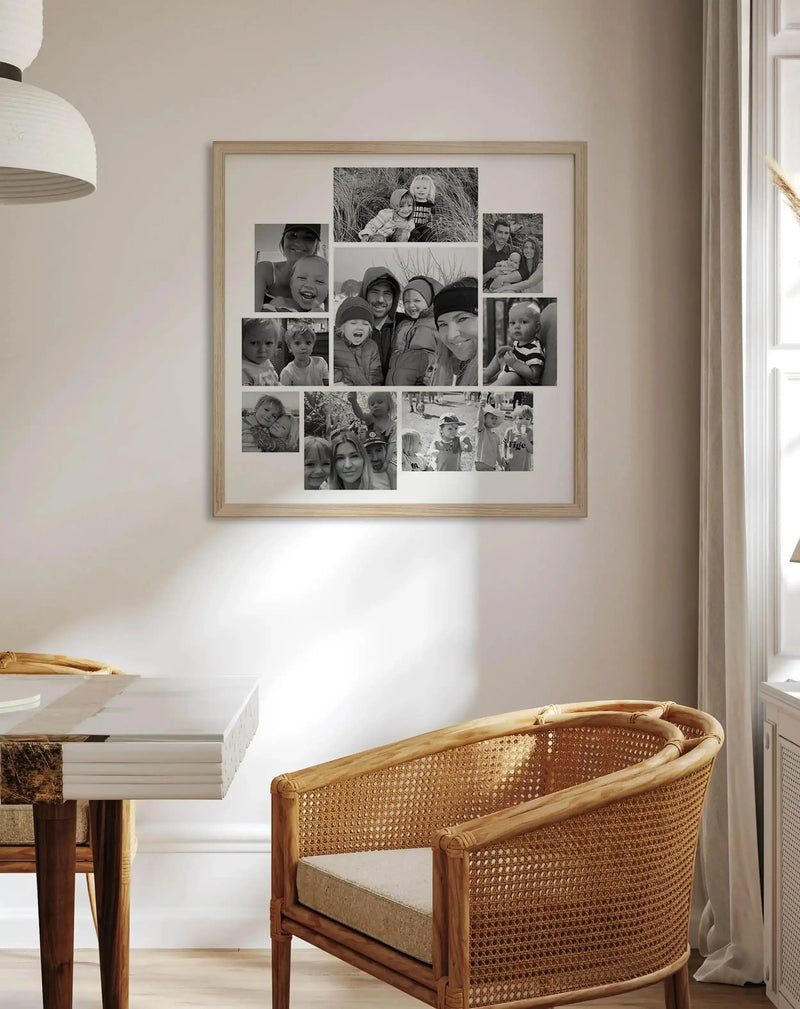 Lifestyle shot of a dining room featuring a large framed art print on a light beige wall. The print is a collage of 9 black and white family photographs, displayed in a natural wood frame with a white border. The room includes a white marble-topped table with dark wood legs and a light wood and woven cane chair with a beige cushion. A white pendant light hangs in the upper left, and a window with a sheer curtain is on the right.