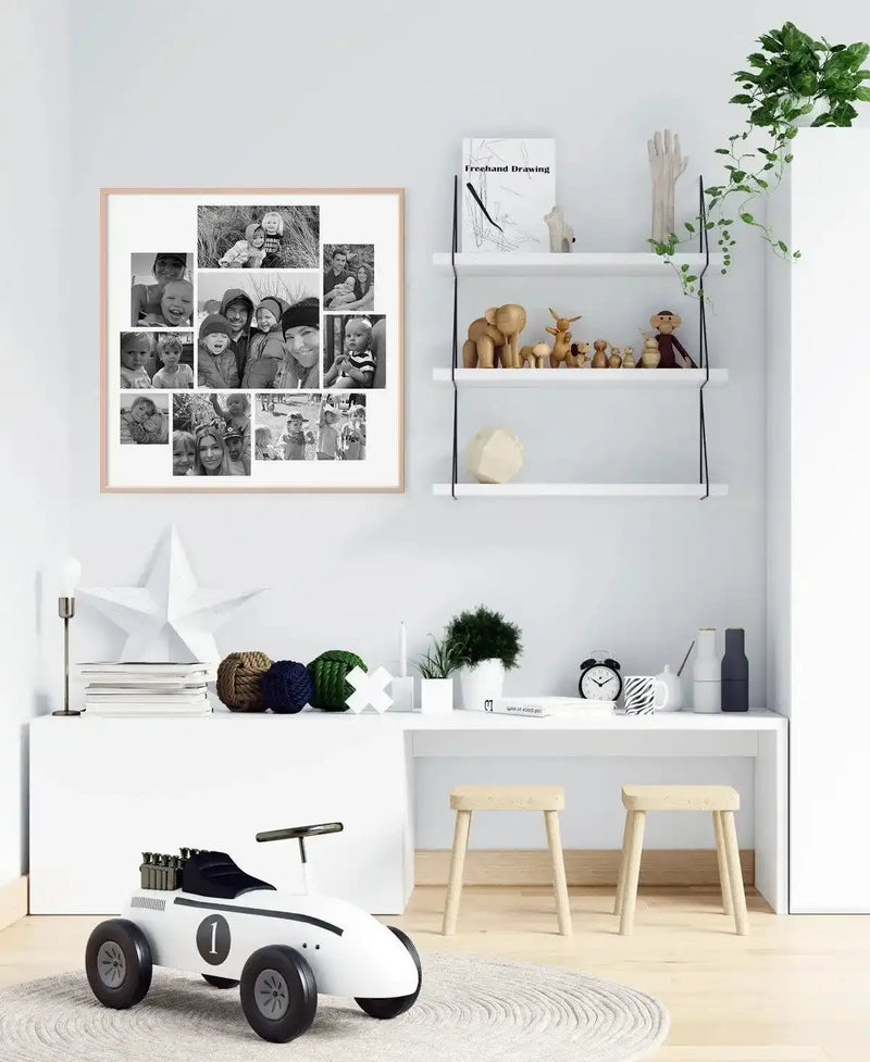 Lifestyle shot of a child's room featuring a framed art print on a white wall above a white console table. The print is a black and white photo collage of 9 square photographs, each depicting families and children, with a natural wood frame and white border. The room also includes white shelves with wooden toys, a white desk with two wooden stools, and a white toy race car on a round rug.