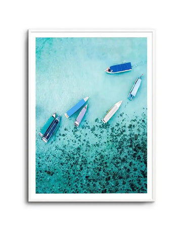 Framed art print featuring an aerial photograph of six fishing boats scattered across shallow turquoise water. The boats display blue, white, and purple hulls with varying orientations. The upper portion shows clear, pale turquoise water, while the lower half transitions to deeper teal with dark green seagrass or coral visible beneath the surface. The photograph is mounted in a white frame with white border, creating a serene coastal scene with natural depth and color gradation.