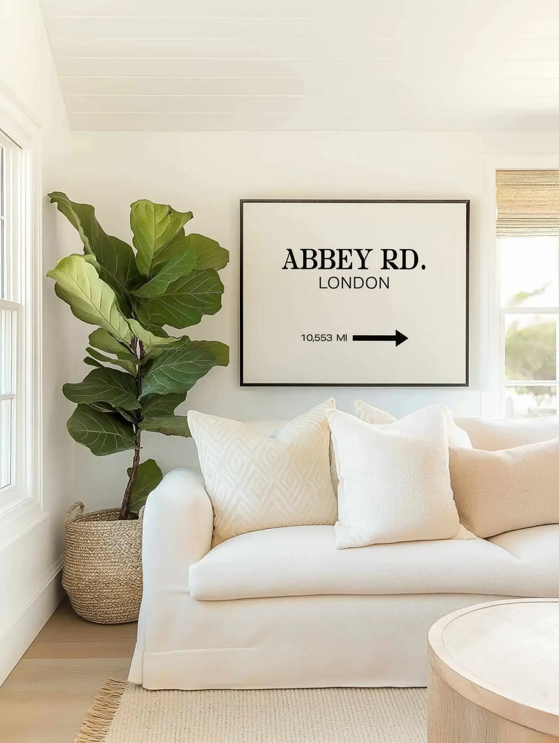 Lifestyle shot of a minimalist living room featuring a large framed art print displayed above a curved white sofa. The print features the text "ABBEY RD. LONDON" in black serif font, with "10,553 MI" and a black arrow pointing right below it, all on a white background, with a black frame and white border. The room includes a large fiddle-leaf fig plant in a woven basket, cream-colored throw pillows, and a light wood coffee table.