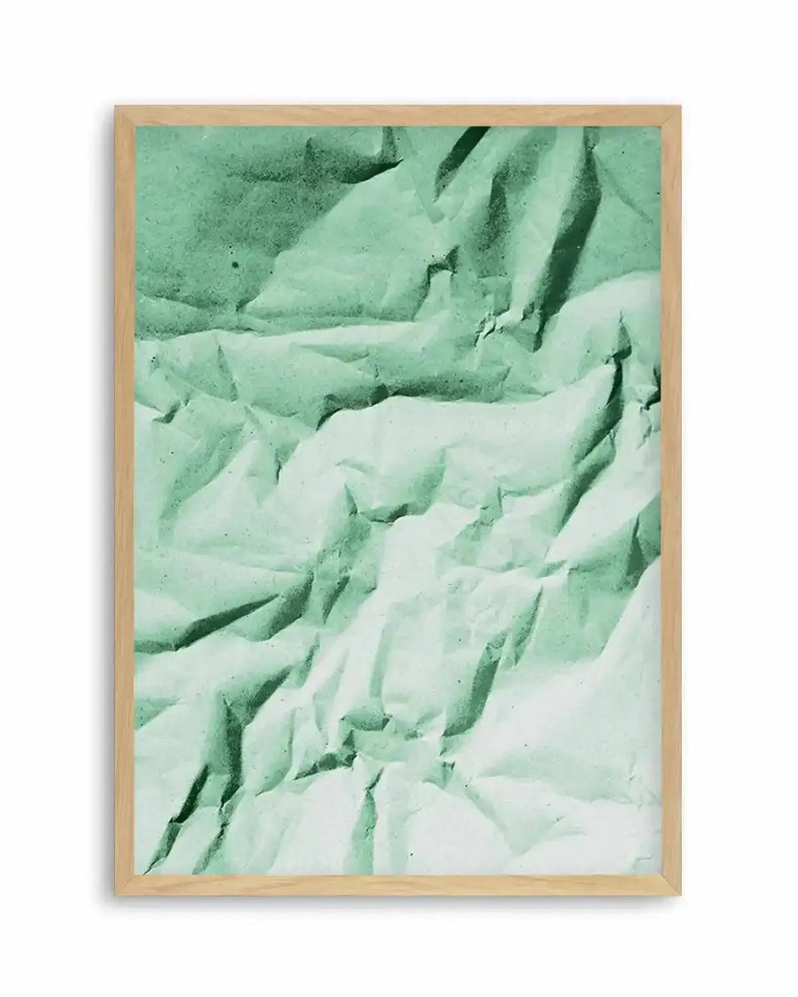 Abstract art print featuring a crumpled sheet of paper in varying shades of mint green, sage green, and pale green, creating deep shadows and bright highlights that emphasize the texture and folds. Printed on fine art paper and displayed in a natural oak frame with white border.