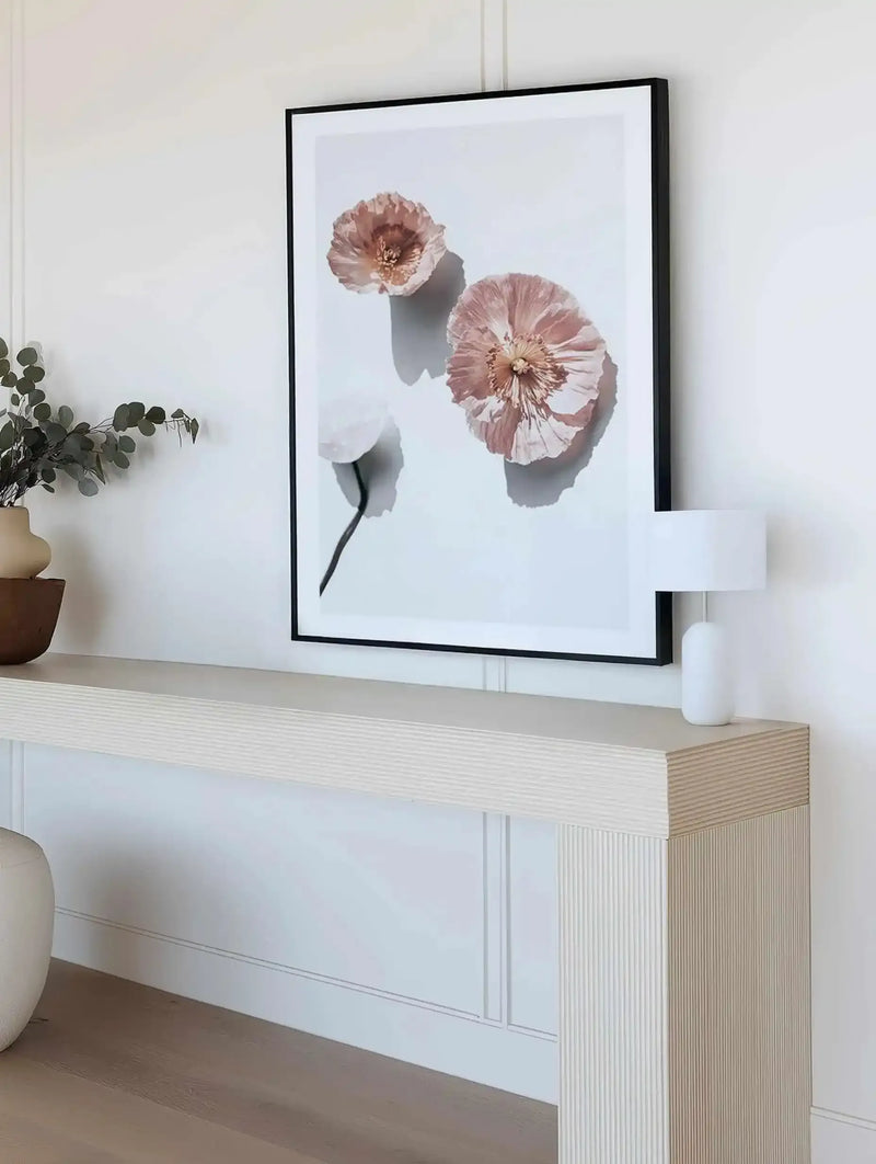 Lifestyle shot of a minimalist entryway featuring a large framed art print displayed above a fluted console table. The print features three abstract poppies with ruffled petals in shades of dusty rose and cream against a bright white background, with a black frame and white border. A small white lamp sits on the console table, and a vase with eucalyptus branches is visible to the left.