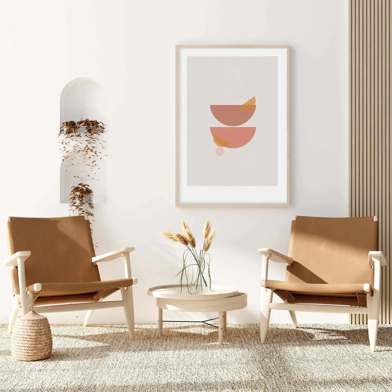 Lifestyle shot of a minimalist living room featuring the Abstract Sienna II art print displayed above a round wooden coffee table. The print features two abstract half-circle shapes in sienna and dusty rose with small ochre and blush pink accents, set against a light grey background with a natural wood frame and white border. Two light wood and tan leather armchairs flank the table, and a woven basket sits on a textured cream rug.