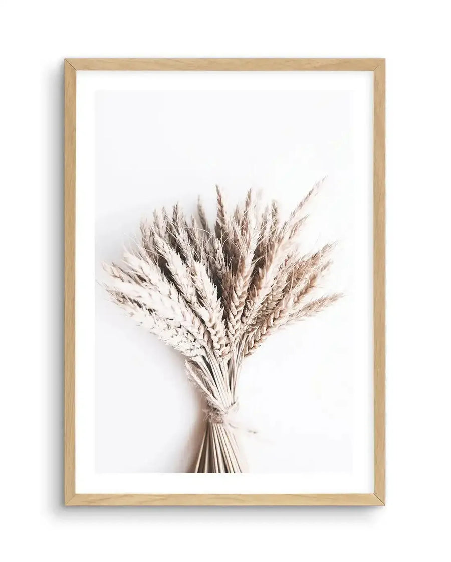 Framed photography art print featuring a close-up of a bouquet of dried wheat stalks in various shades of beige and light brown, tied together with twine, against a clean white background. The natural wood frame includes a white border.