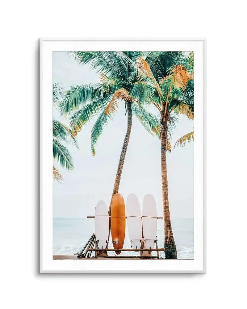 Framed art print featuring a photograph of surfboards stacked on a wooden rack beneath two tall palm trees on a tropical beach. An orange surfboard is prominently centered among white surfboards. The ocean and overcast sky are visible in the background. The image has soft, muted tones with green palm fronds, tan tree trunks, and pale blue-gray sky. The photograph is matted with a white border and displayed in a light gray frame.