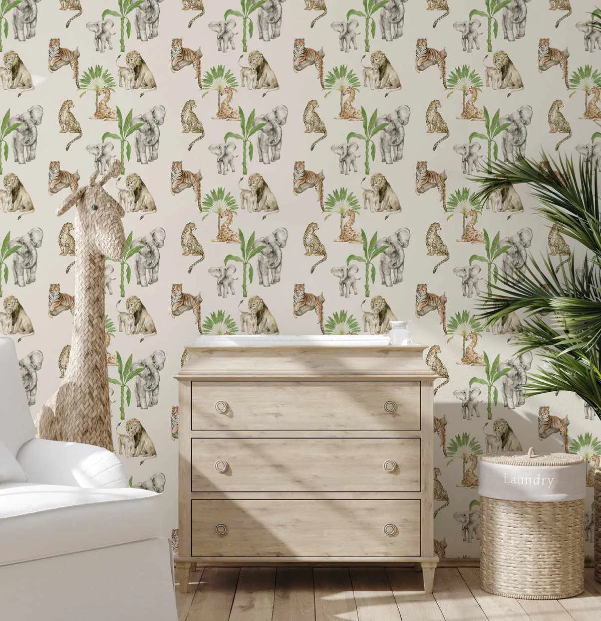 Lifestyle shot of a nursery featuring African animals wallpaper. The wallpaper has a repeating pattern of hand-painted safari animals including elephants, lions, tigers, cheetahs, and giraffes, alongside palm trees and green foliage on a cream background. A light wood changing table with three drawers is centered against the wall, with a white armchair and a large woven giraffe toy on the left, and a tall green plant and a woven laundry basket on the right.