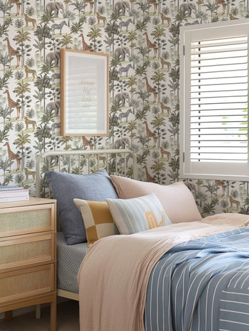 "African Adventure wallpaper in a child's bedroom featuring a repeating pattern of hand-drawn safari animals including giraffes, elephants, zebras, and leopards amidst lush green and brown palm trees and tropical foliage on a white background. The wallpaper is visible behind a white metal bed frame with blue and pink bedding, a natural wood bedside table with rattan drawers, and a framed print that reads 'dream big little one'."