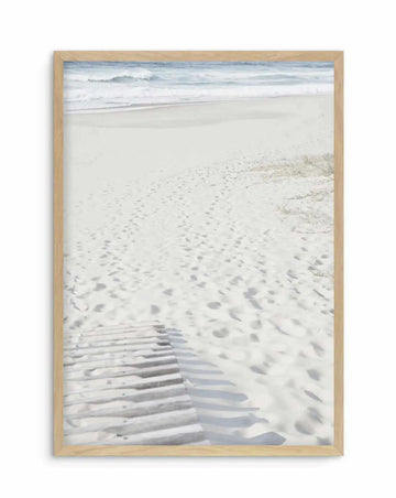 Framed photography art print of a white sand beach with a wooden boardwalk leading towards the ocean, displayed in a natural oak frame with white border. The composition features gentle blue waves breaking on the shore under a bright sky, with numerous footprints visible in the sand.