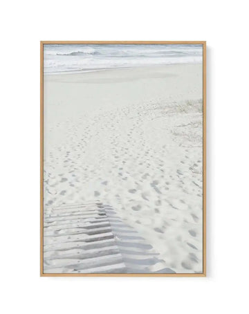 Framed canvas art print with natural wood frame featuring a serene beach scene. The composition shows footprints and tire tracks in pale sand in the foreground, with gentle waves and white foam in the middle distance, and a misty gray-blue ocean with distant hills under an overcast sky in the background. The photograph has a soft, muted color palette of cream, pale gray, and cool blue tones, creating a calm, contemplative coastal mood.