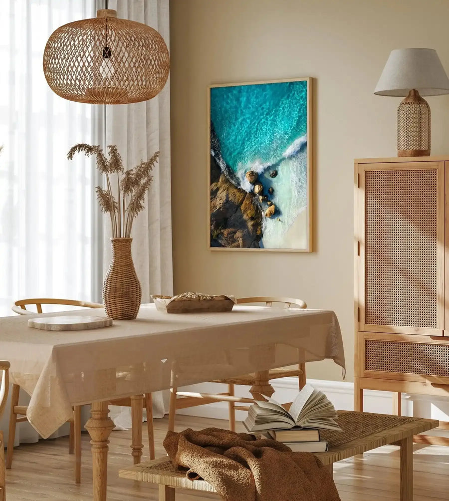 Lifestyle shot of a dining room featuring a framed art print of an aerial beach scene with natural wood frame and white border. The print depicts turquoise ocean water meeting a sandy beach with rocky outcrops, and a white wave breaking on the shore. The room includes a light wood dining table with a beige linen tablecloth, a woven pendant light, a tall wicker vase with dried pampas grass, and a cane cabinet.