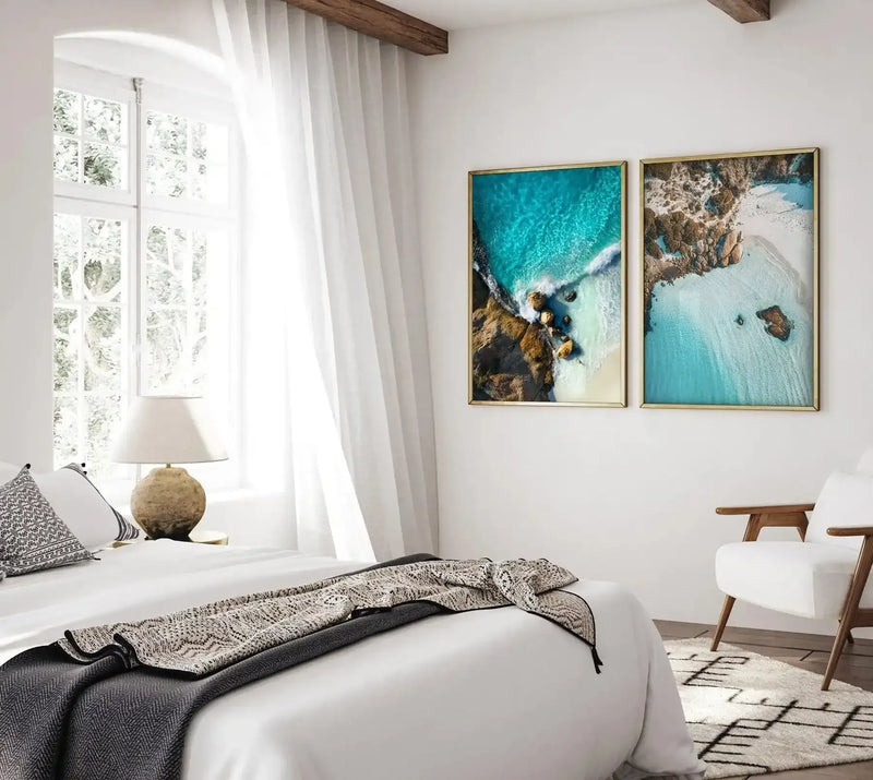 Lifestyle shot of a bright bedroom featuring two framed art prints above a white armchair. The prints are aerial photographs of a coastline with turquoise water, white sand, and rocky outcrops, displayed in gold frames. The room has a large window with white sheer curtains, a bed with white linen and patterned throws, and a round textured lamp.