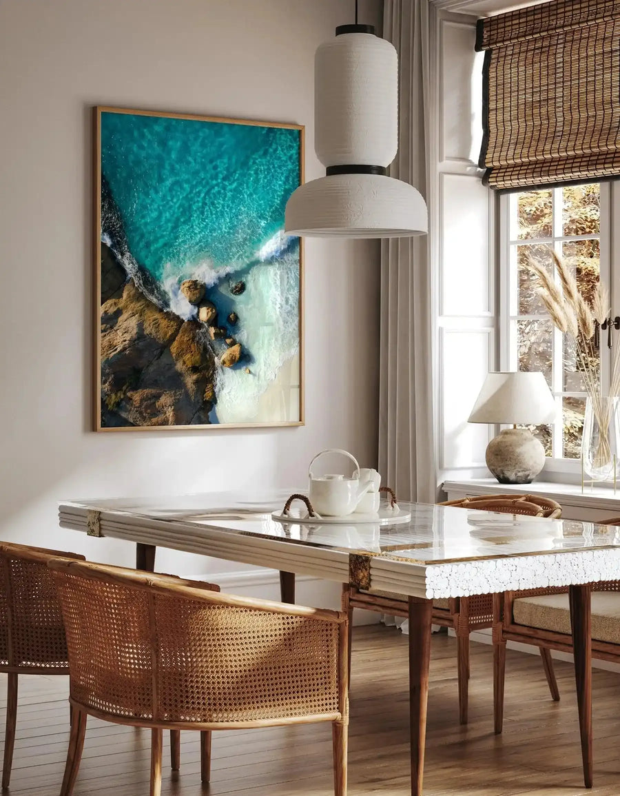 Lifestyle shot of a dining room featuring a large framed art print of an aerial beach scene. The print, displayed in a natural wood frame with a white border, captures turquoise ocean water meeting a sandy beach and rocky coastline. The dining room includes a long table with a clear top and white textured sides, wicker chairs, a white pendant light, and a window with a woven blind.
