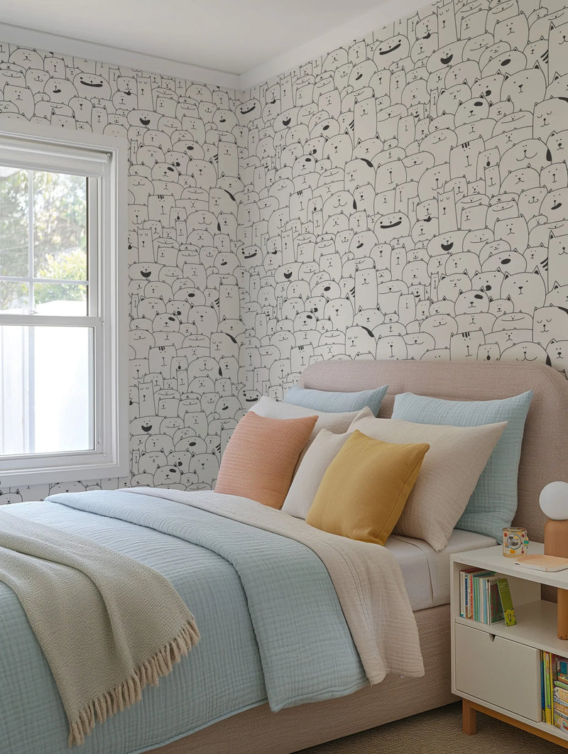 Lifestyle shot of a bedroom featuring "All the Cats" wallpaper. The wallpaper covers two walls and features a repeating pattern of hand-drawn, cartoon-style cats in black outline on a white background, with each cat having a unique expression and shape. The room includes a bed with a light beige upholstered headboard, light blue bedding, and an assortment of pillows in peach, cream, and yellow. A white nightstand with books and a lamp is visible on the right.