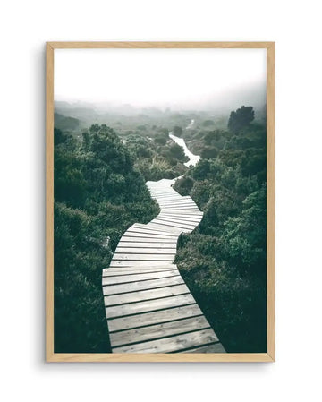 Framed photography art print featuring a winding wooden boardwalk through a misty, lush green landscape, displayed in a natural oak frame with white border. The path curves through dense, dark green foliage under an overcast, bright white sky, leading towards a distant, hazy horizon.