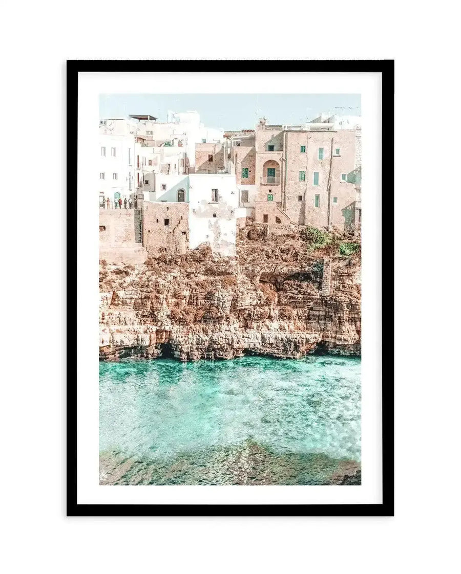 Framed photography art print of a coastal town built into a cliffside overlooking turquoise water, with a black frame and white border. The composition features white and light brown buildings with green window shutters, perched on rugged brown and tan rock formations, with clear, shallow blue-green water visible below.
