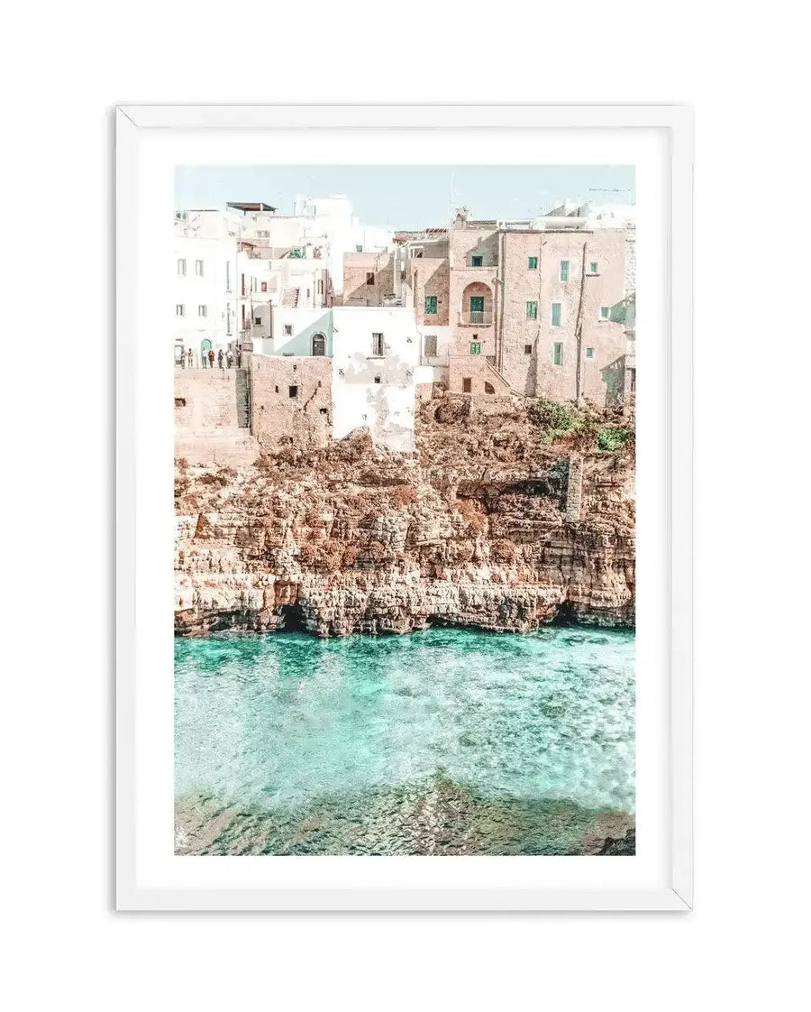 Framed photography art print of a coastal town built into a cliffside above turquoise water, with a white frame and white border. The composition features buildings in shades of white and pale terracotta with green window shutters, perched on a rugged brown and orange cliff face. Below the cliff, the clear, shallow water reveals the rocky seabed.