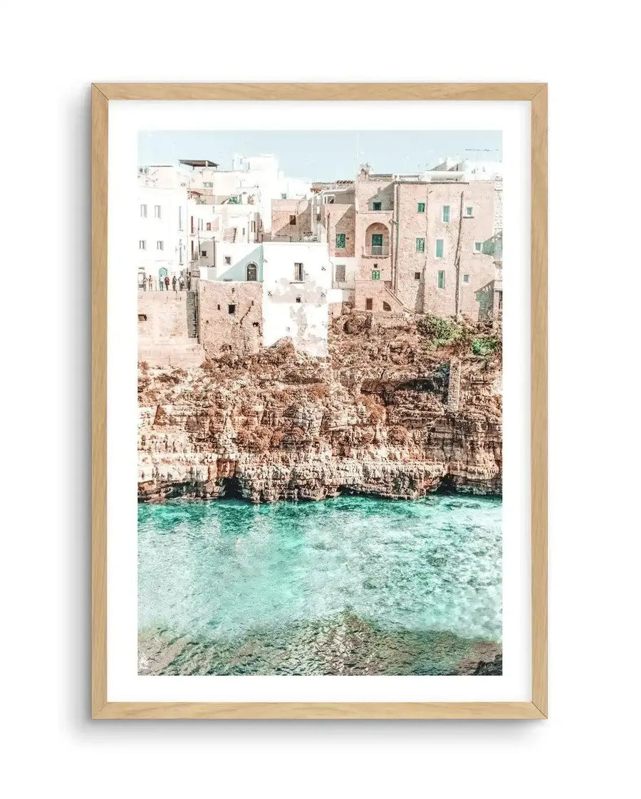 Framed photography art print of a coastal town built into a cliffside above clear turquoise water, with natural wood frame and white border. The composition features white and pale pink buildings with green shutters, perched on a rugged brown and orange rock face, overlooking a calm, shallow sea with visible underwater rock formations.