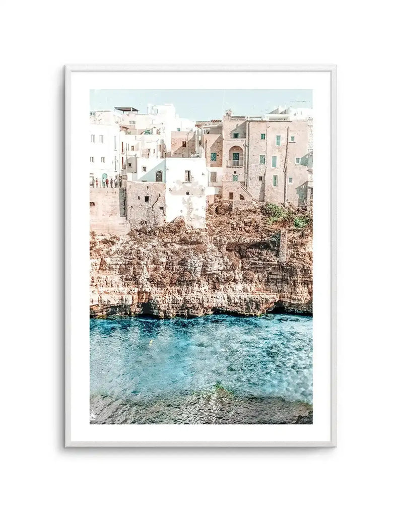 Framed photography art print of a coastal town built into a cliffside overlooking clear blue water, with a white frame and white border. The composition features white and beige buildings with green shutters on a rocky cliff, with the turquoise sea below.