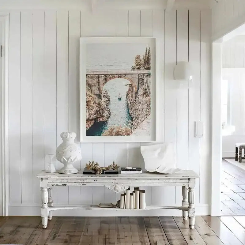 Lifestyle shot of a white entryway with a large framed art print displayed above a distressed white console table. The art print is a photograph of the Amalfi Coast, featuring a stone bridge spanning a deep gorge with a narrow river flowing through it, and a small boat on the water. The room has white vertical paneling on the walls and light wood flooring. The console table is adorned with white decorative vases, dried botanical elements, and books.