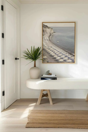 Lifestyle shot of a minimalist entryway featuring the Amalfi Coast Life I framed canvas art print displayed above a curved white bench. The print is a photograph of a sandy beach with rows of straw umbrellas and lounge chairs, a calm blue ocean, and a white cliff face, framed in natural wood with a white border. A tall green plant in a light-colored vase sits on the bench, next to a stack of books and decorative objects. The room has light wood floors and a natural fiber rug.