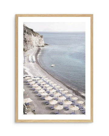 Framed photography art print of a serene coastal beach scene with a natural wood frame and white border. The composition features a long stretch of pebble beach lined with rows of straw beach umbrellas and sun loungers, with a white cliff face on the left and calm blue-grey ocean extending to the horizon. A small white boat floats in the water near the shore.