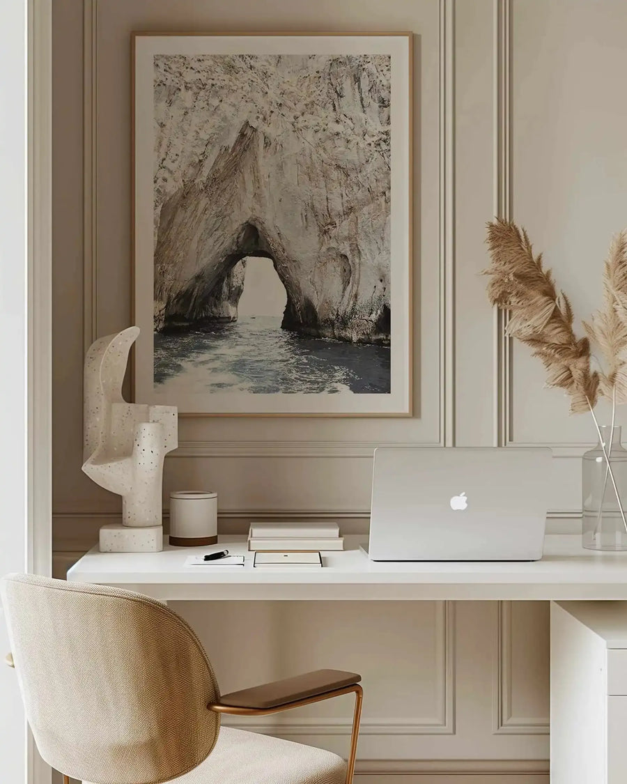 Lifestyle shot of a minimalist home office with a framed art print of the Amalfi Coast displayed above a white desk. The print features a natural rock archway over deep blue ocean water, with a small boat visible in the distance, framed in natural wood with a white border. The desk holds a white laptop, a sculptural white speckled art piece, and dried pampas grass in a clear vase.