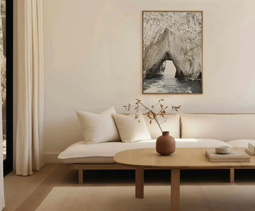 Large framed canvas art print displayed above a curved white sofa in a minimalist living room. The print features a natural rock archway over calm ocean water, with a small boat visible in the distance, in muted grey, white, and blue tones, with a natural wood frame. The room includes cream pillows, a light wood coffee table with a terracotta vase holding dried leaves, and a cream rug.