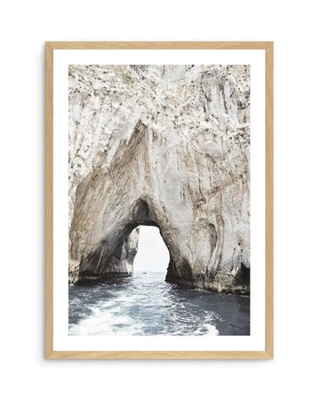 Framed photograph of a natural rock archway over calm ocean water, with a natural wood frame and white border. The composition features a large, light-colored rock formation with a prominent arch in the center, revealing a glimpse of the open sea and a small boat in the distance. The water in the foreground shows gentle ripples and white foam.