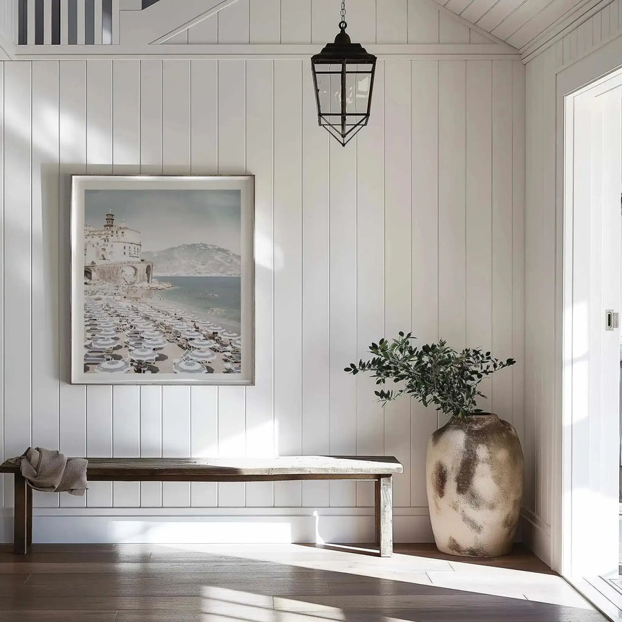 Lifestyle shot of a hallway featuring a large framed art print of the Amalfi Coast. The print, displayed in a natural wood frame with a white border, depicts a vibrant beach scene with numerous striped umbrellas, a white building on a cliff, and mountains in the background. The hallway has white vertical paneling, a dark wooden bench with a draped throw, and a large textured vase with green foliage. Sunlight streams across the wooden floor.
