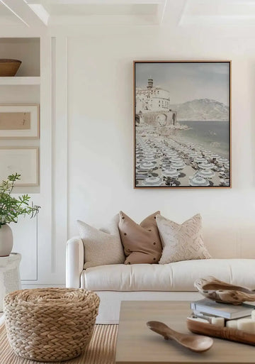 Lifestyle shot of a minimalist living room featuring the Amalfi Coast Life III framed canvas art print displayed above a cream sofa. The print is a photograph of a bustling beach scene with rows of striped umbrellas and sunbathers, a historic white building with a bell tower on a cliff, and calm blue water with distant mountains under a pale sky, framed in natural wood. The sofa is adorned with textured cream and brown pillows, and a woven basket sits on a striped rug in the foreground.