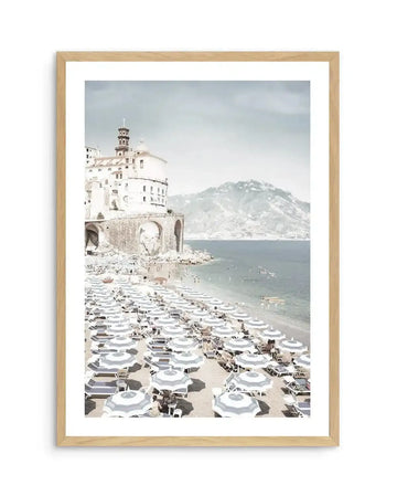 Framed photography art print of a coastal beach scene in Amalfi, Italy, with a natural wood frame and white border. The composition features a white multi-story building with a bell tower on a stone archway overlooking a beach filled with rows of striped grey and white umbrellas and sun loungers. The calm blue sea stretches to a distant mountain range under a pale grey sky.