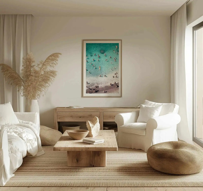 Lifestyle shot of a minimalist living room featuring the Amalfi Days art print displayed above a wooden console table. The print is a framed aerial photograph of a beach with turquoise water and people swimming and relaxing on the sand, with a natural wood frame and white border. The room has light cream walls, a white sofa, a large vase of dried pampas grass, and a woven rug.