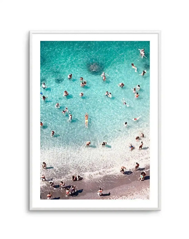 Framed photography art print featuring an aerial view of a beach scene with a white frame and white border. The composition shows numerous people swimming in clear turquoise water and walking on the light brown sand at the water's edge, with gentle waves breaking on the shore.