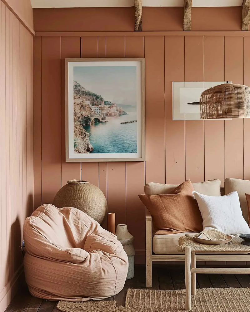 Lifestyle shot of a living room with a large framed art print of a coastal town displayed on a terracotta-colored paneled wall. The print, titled "Amalfi Sunsets I," features a picturesque view of white buildings nestled into a rocky cliffside overlooking a calm blue sea, with a stone bridge and a distant breakwater. The room includes a light pink bean bag chair, a woven basket, and a light wood sofa with cream and rust-colored cushions, and a woven pendant light.