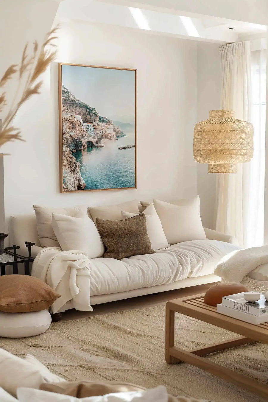 Large framed canvas art print displayed above a cream-coloured sofa in a bright, minimalist living room. The print features a photograph of a coastal town built into cliffs overlooking a calm, turquoise sea, with a bridge extending over the water. The print has a natural wood frame. The room includes a woven pendant light, a light wood coffee table, and various cream and brown cushions.