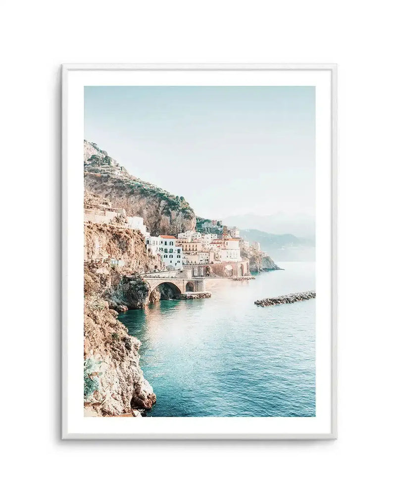 Framed photography art print of a coastal town built into a cliffside, with a white frame and white border. The scene features white and terracotta buildings nestled into a rocky, green-covered cliff that slopes down to a calm, turquoise-blue sea. A stone bridge with arches spans a small bay, and a breakwater extends into the water. The sky is a clear, pale blue, creating a serene and bright atmosphere.