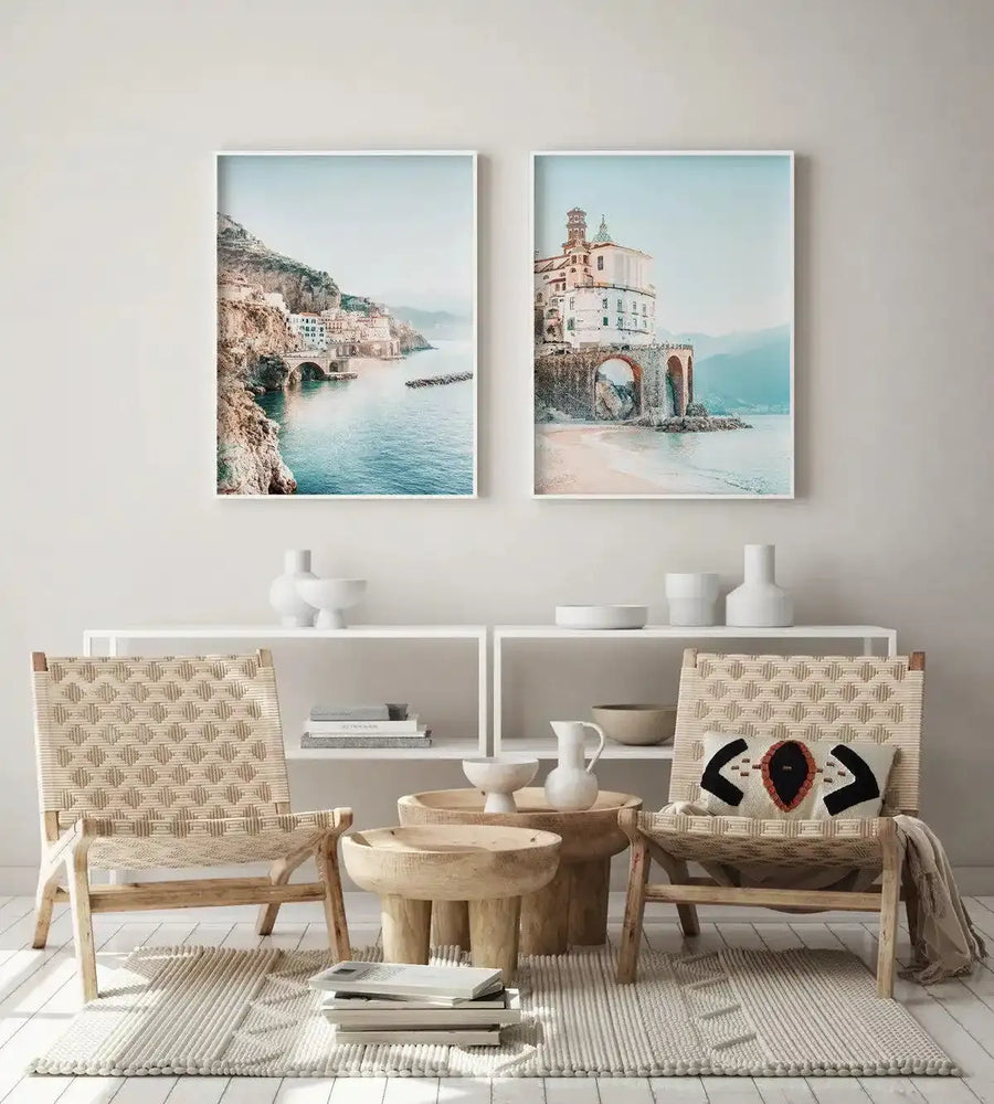 Two framed photography art prints displayed side-by-side above a white shelf in a minimalist living room. The left print features a coastal town built into a cliffside overlooking turquoise water, with a bridge connecting parts of the town. The right print shows a large white building with a bell tower on a stone archway overlooking a sandy beach and turquoise water. The room includes two woven chairs, wooden side tables, and white ceramic vases.