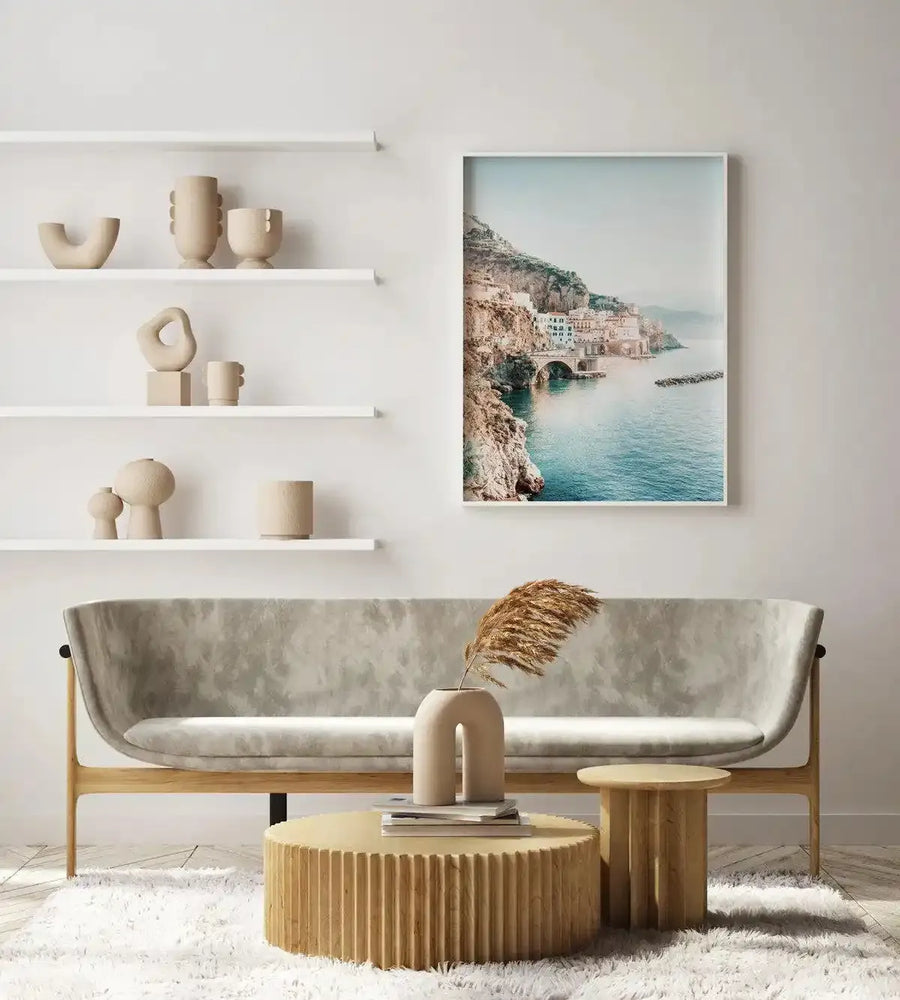 Large framed art print displayed above a curved light grey sofa in a minimalist living room. The print is a photograph of a coastal town built into cliffs overlooking the ocean, with a bridge connecting parts of the town over the water, and a light blue sky. The print has a white frame and white border. The room includes white floating shelves with various beige ceramic vases and sculptures, a round fluted light wood coffee table with books and a beige vase holding dried pampas grass, and a white shag rug.