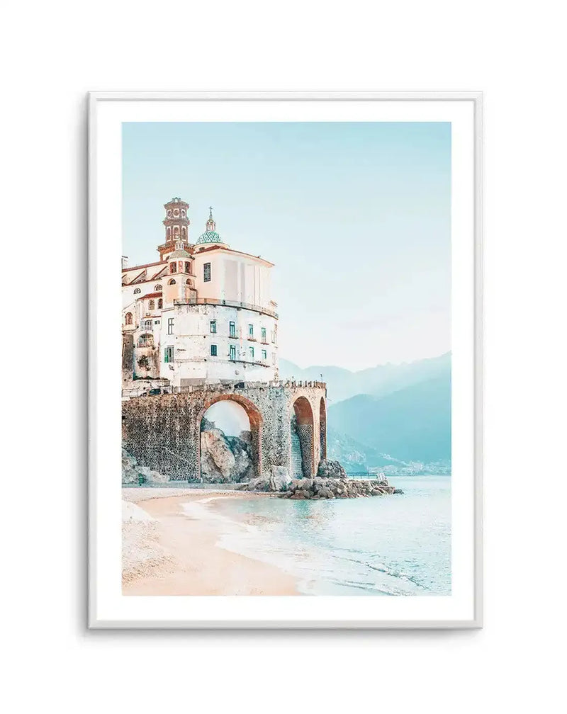 Framed photography art print of a coastal town in Italy, with a white building featuring a green dome and bell tower built on a stone archway over a sandy beach. The sky and ocean are a soft pastel blue, and distant mountains are visible in the background. The print is displayed in a white frame with a white border.