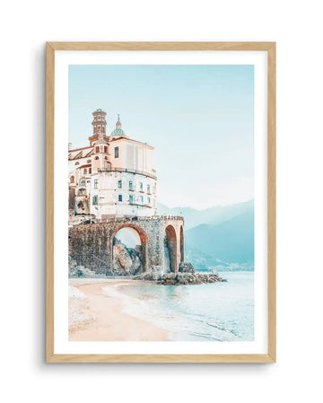 Framed photography art print of a coastal town with a natural wood frame and white border. The composition features a white building with a green dome and bell tower perched on a stone wall with arched openings, overlooking a sandy beach and calm turquoise sea. Distant mountains are visible under a pale blue sky.