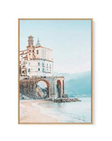 Framed canvas art print depicting a cream and terracotta Italian coastal church perched on a rocky stone archway overlooking a turquoise sea and sandy beach, with misty mountains in the background under a pale blue sky. The artwork features soft, muted tones in a painterly style with warm peachy hues on the architecture and cool aquamarine water. The canvas is mounted in a natural wood frame with a white border.