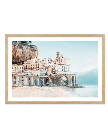 Framed photography art print of a coastal town built into a cliffside with a natural oak frame and white border. The composition features white and cream buildings with terracotta roofs, some supported by stone arches, overlooking a sandy beach and calm turquoise sea under a bright, clear sky.