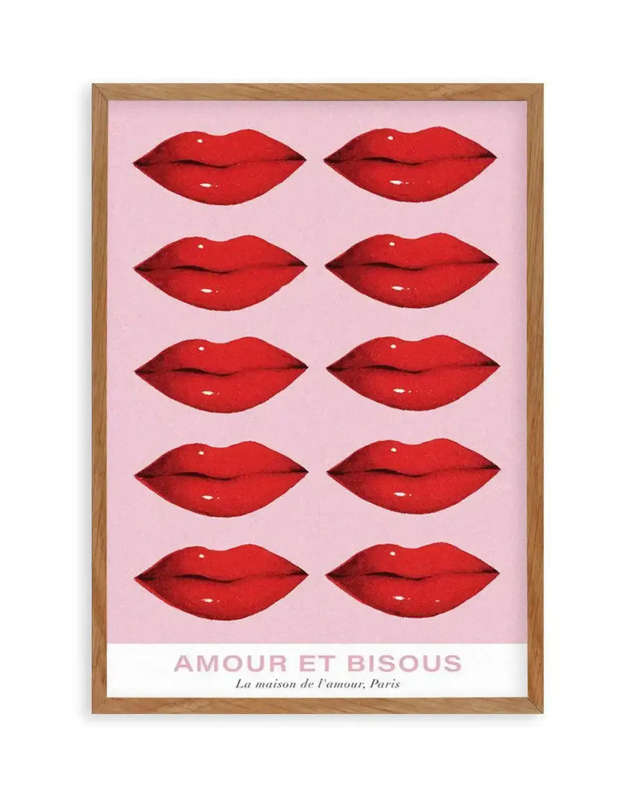Art print featuring ten glossy red lips arranged in five rows of two on a pale pink background, displayed in a natural oak frame with white border. The bottom of the print features the text "AMOUR ET BISOUS" in pink capital letters, with "La maison de l'amour, Paris" in smaller pink script below.