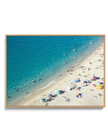 "Framed canvas print of an aerial photograph of a vibrant beach scene with a natural wood frame. The composition features clear turquoise water with swimmers, a sandy beach dotted with colourful umbrellas in shades of blue, green, yellow, and pink, and sunbathers relaxing on towels."